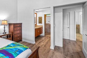 A hotel room with a bed, a dresser, a nightstand, a lamp, a bathroom with a sink and mirror, and a door leading to another room. - Wailea Elua 1104 - Ocean Views, Central A/C, Walk to Ulua Beach, Shops & Dining! (Kihei)