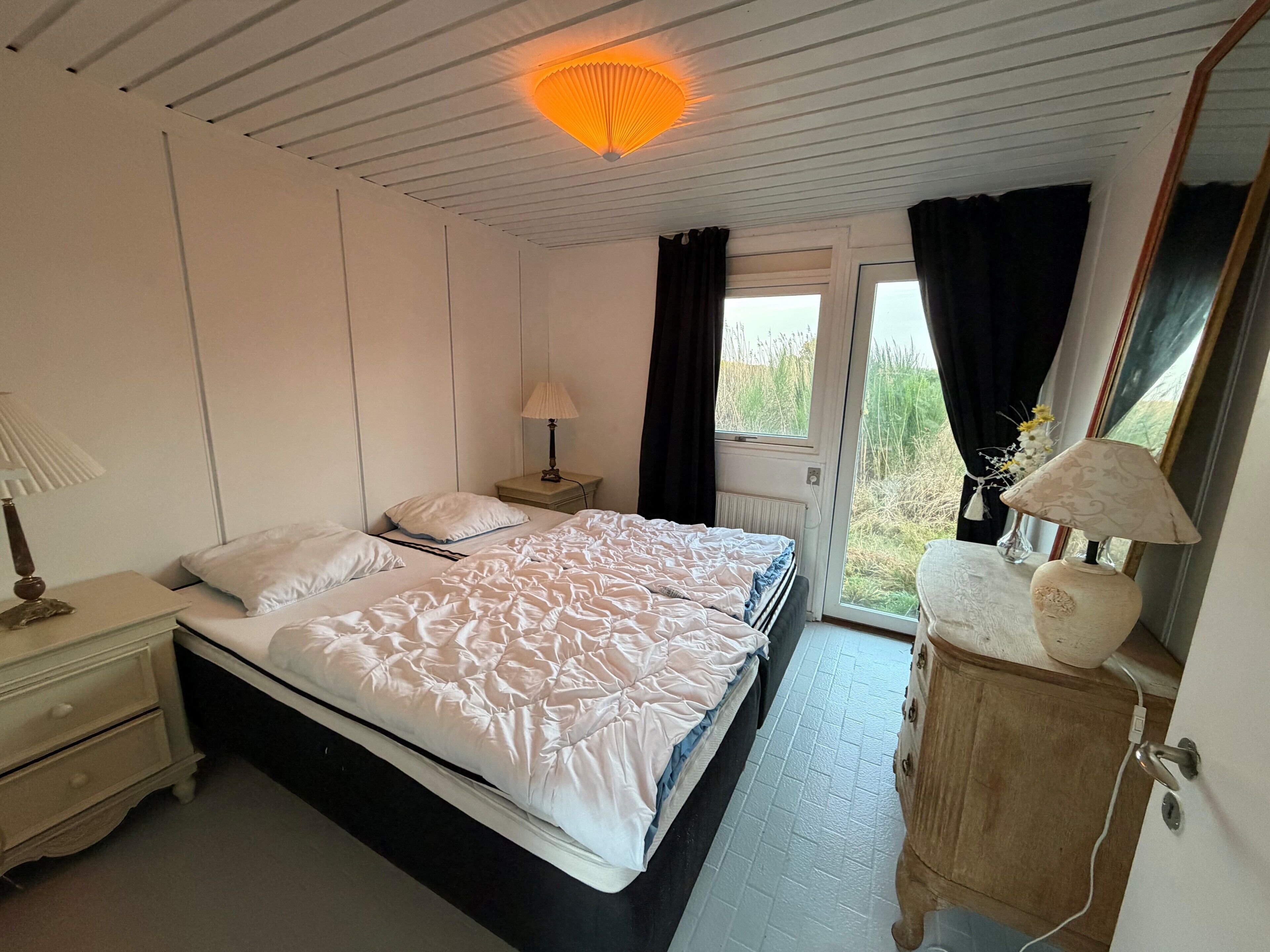 Een slaapkamer met een bed, nachtkastjes, een lamp, een kaptafel en een raam met gordijnen.