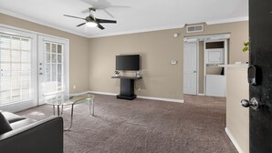 A living room with a ceiling fan, a flat-screen TV on a stand, a glass coffee table, and a door leading to a kitchen. - Kingsgate One Bedroom Stay (Beaumont)