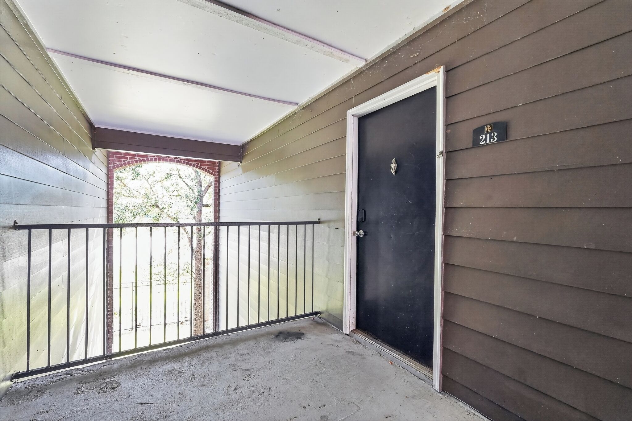 Puerta cerrada con placa, balcón con barandilla metálica y vista a un árbol a través de una ventana.
