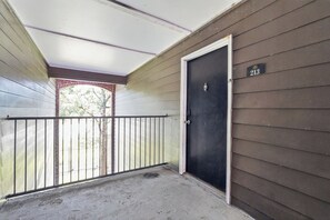 Une porte fermée avec une plaque numérotée, un balcon doté d’une rambarde en métal et une vue sur un arbre à travers une fenêtre.