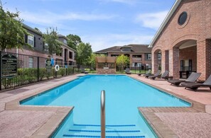 Ein rechteckiger Pool mit einer Metallleiter, umgeben von einem Backsteinhaus mit einer überdachten Terrasse und Liegestühlen.