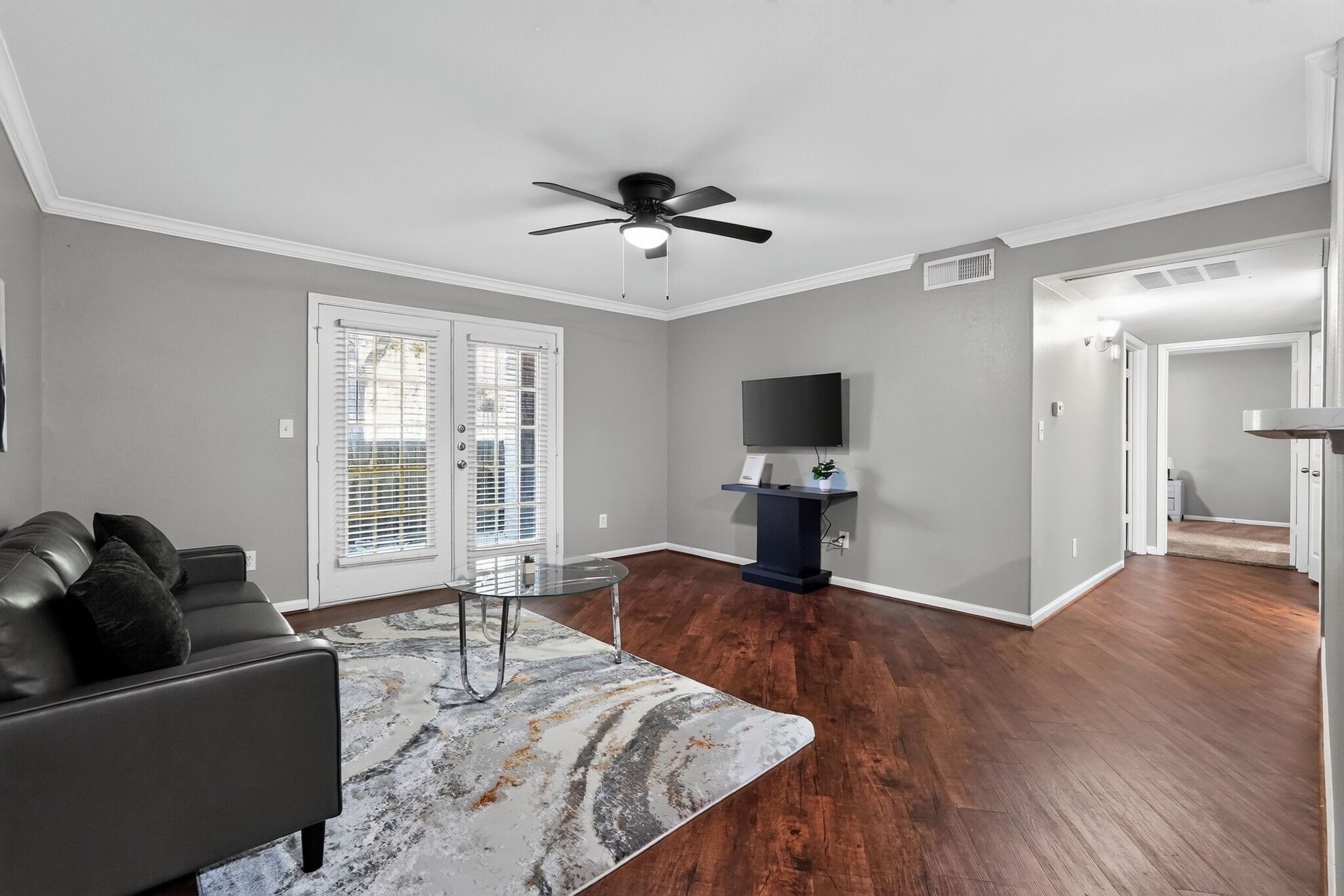 A living room with a sofa, a glass coffee table, a ceiling fan, and a television on a stand.