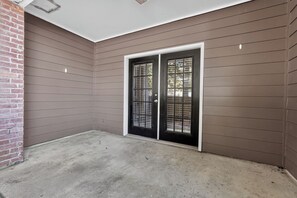 A building exterior with a concrete ground, a brick wall on the left, and a double door with a metal grille. - Comfortable Kingsgate 2BR (Beaumont)