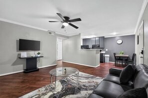 A modern living room with a glass coffee table, a black leather sofa, a ceiling fan, and a flat-screen TV mounted on the wall. - Comfortable Kingsgate 2BR (Beaumont)