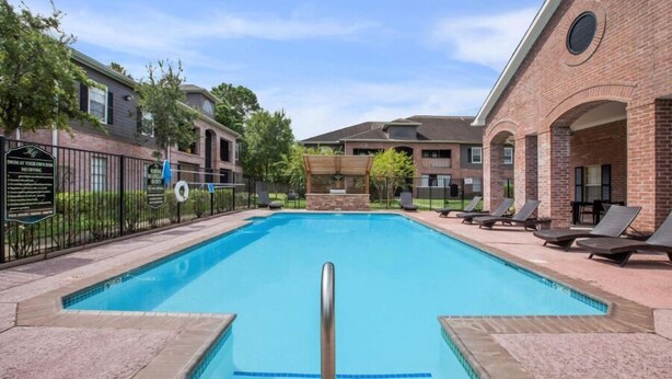 A rectangular swimming pool with a metal ladder, surrounded by a brick building with a covered patio area and lounge chairs. - Relaxing Kingsgate 2BR (Beaumont)