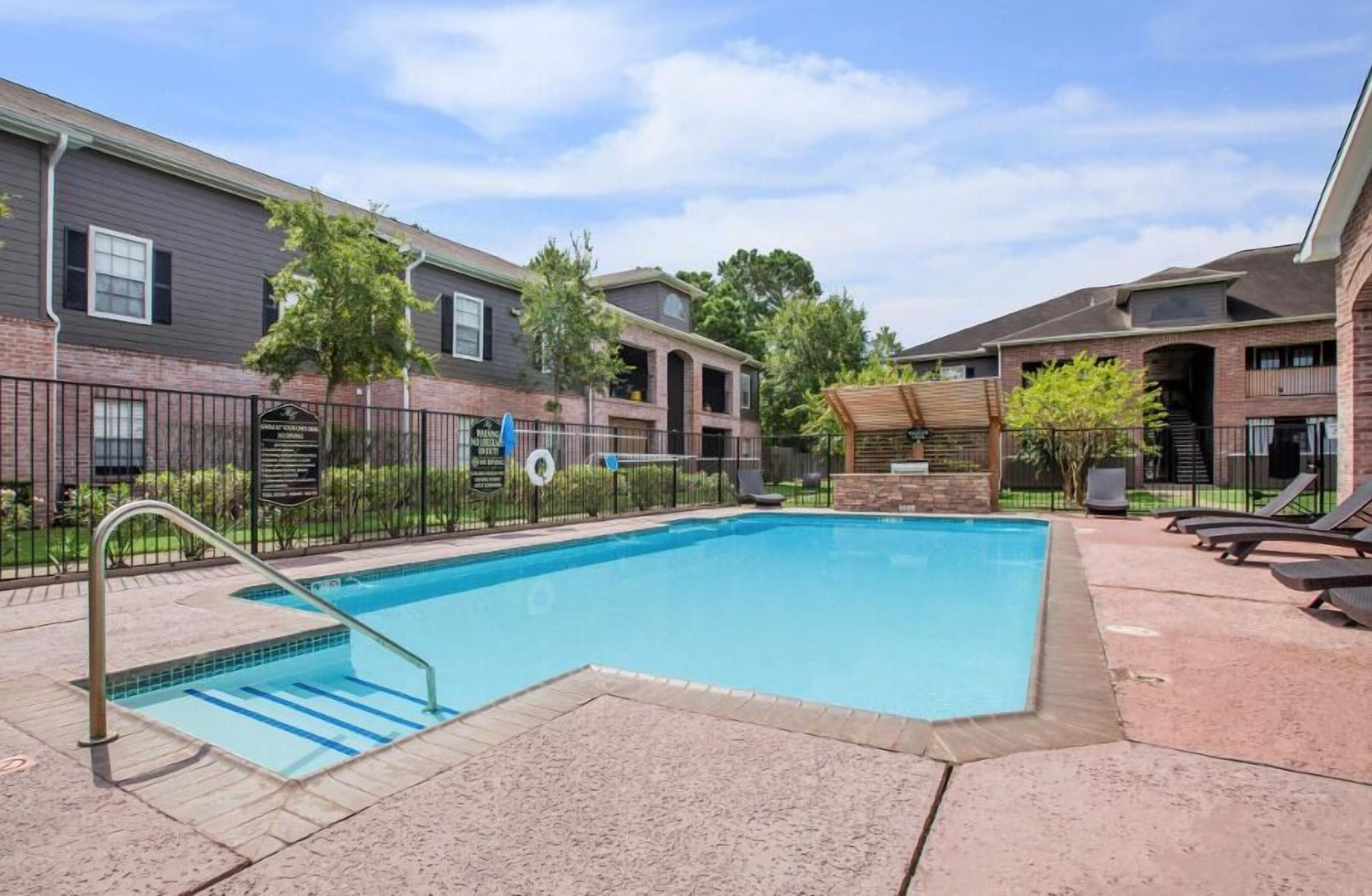 A swimming pool with a metal ladder, surrounded by a brick building with a covered seating area and lounge chairs.