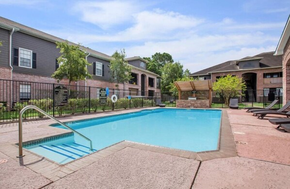 A swimming pool with a metal ladder, surrounded by a brick building with a covered seating area and lounge chairs.