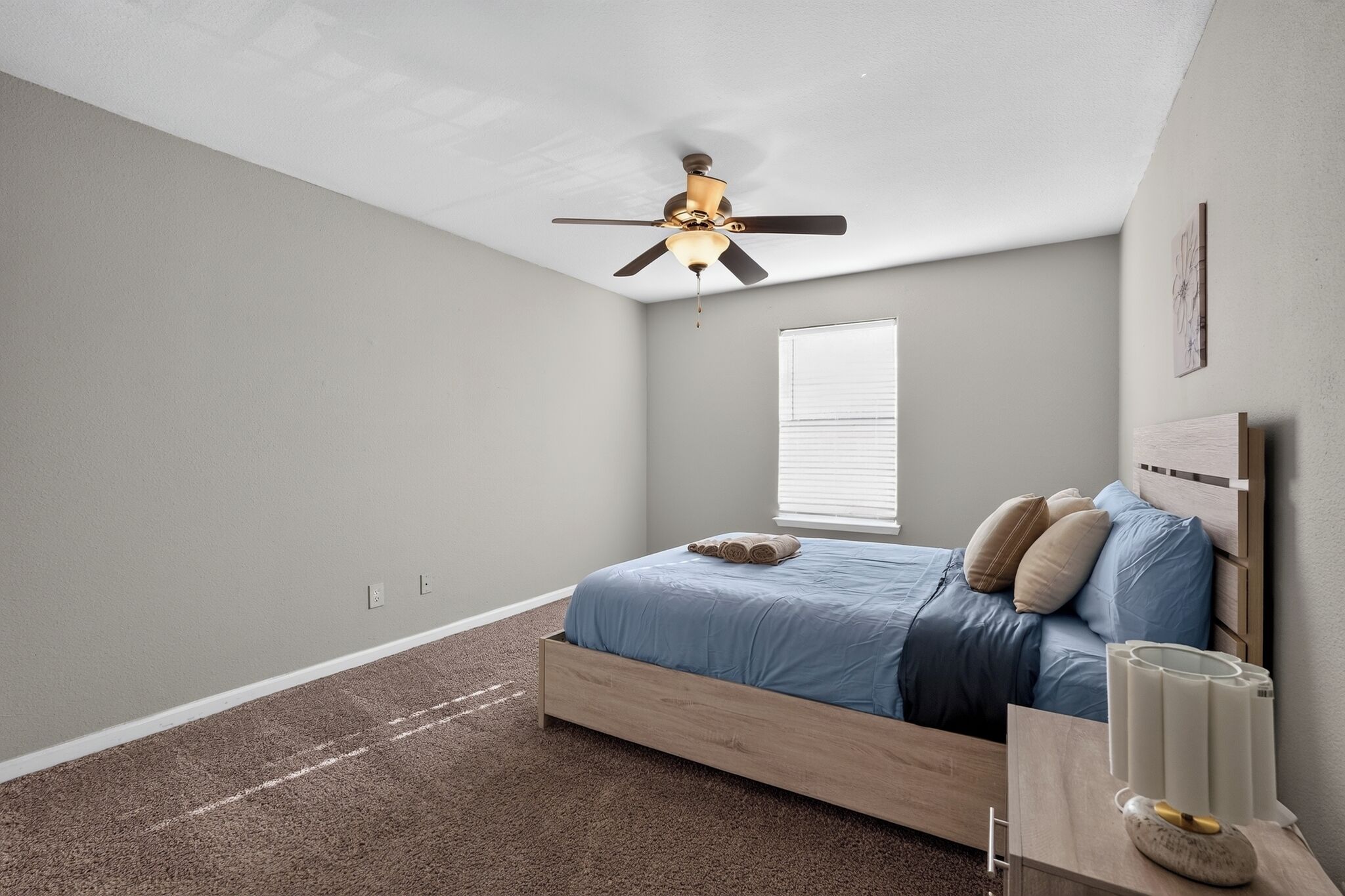 A bedroom with a single bed, a ceiling fan, a window with blinds, and a bedside table with a lamp.