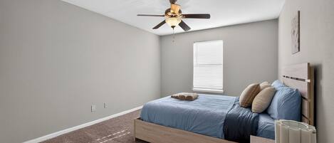 A bedroom with a single bed, a ceiling fan, a window with blinds, and a bedside table with a lamp.