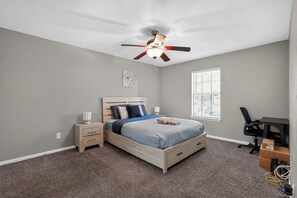 Une chambre à coucher comprenant un lit, des tables de chevet, un bureau, une chaise, un ventilateur de plafond et une fenêtre.