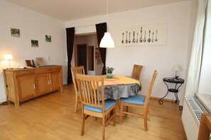 A dining area with a wooden table, chairs, a lamp, and a wall-mounted picture of kitchen utensils. - Appartement im Seedomizil Kummerow (Kummerow)