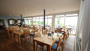 A spacious dining area with wooden tables and chairs, large windows offering a view of the outdoors, and a sign indicating a cocktail price. - Appartement im Seedomizil Kummerow (Kummerow)