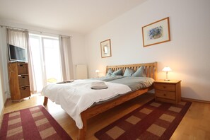 Ein Schlafzimmer mit einem Bett, einem Fernseher, einem Nachttisch mit Lampe, einer Kommode und einem Fenster mit Vorhängen.