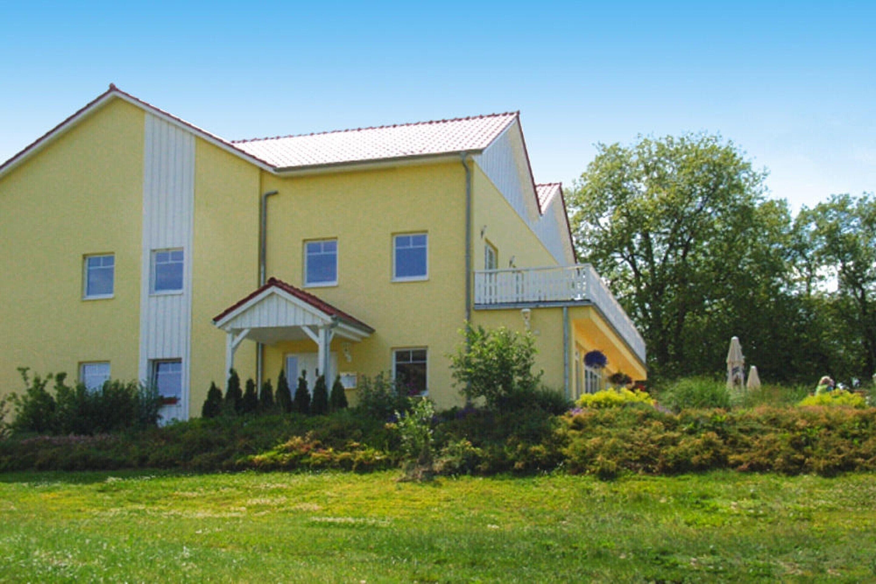 Un immeuble jaune à deux étages, doté d’un balcon au deuxième étage, d’une entrée couverte et d’un jardin en premier plan.