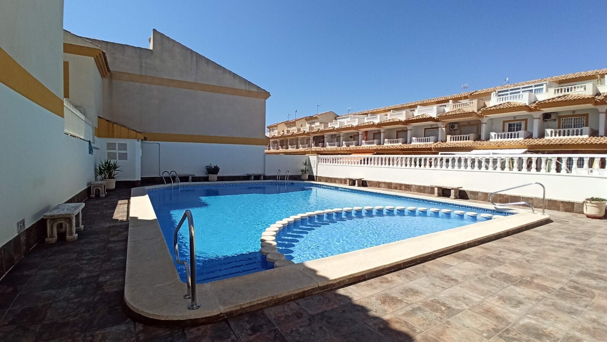 A swimming pool with a curved edge, surrounded by a tiled deck, and a building with balconies in the background.