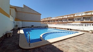 Una piscina de borde curvo, rodeada por una solana con baldosas, y un edificio con balcones al fondo.
