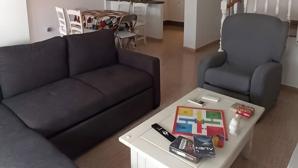 A modern living room with a dark sofa, a white coffee table with remote controls and books, a grey armchair, and a ceiling fan.