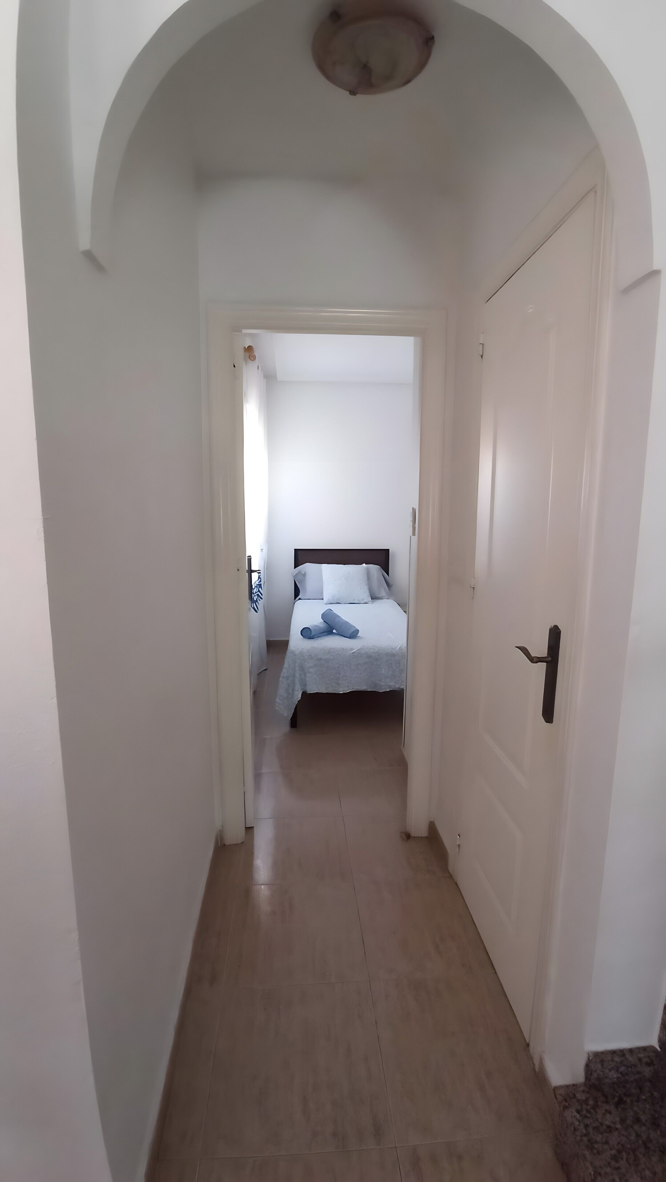 Een gang die leidt naar een slaapkamer met een bed, een deur en een plafondlamp.