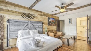 Una camera da letto con una testiera in legno, un letto con lenzuola bianche, una ventola a soffitto, un quadro, una sedia e una porta.