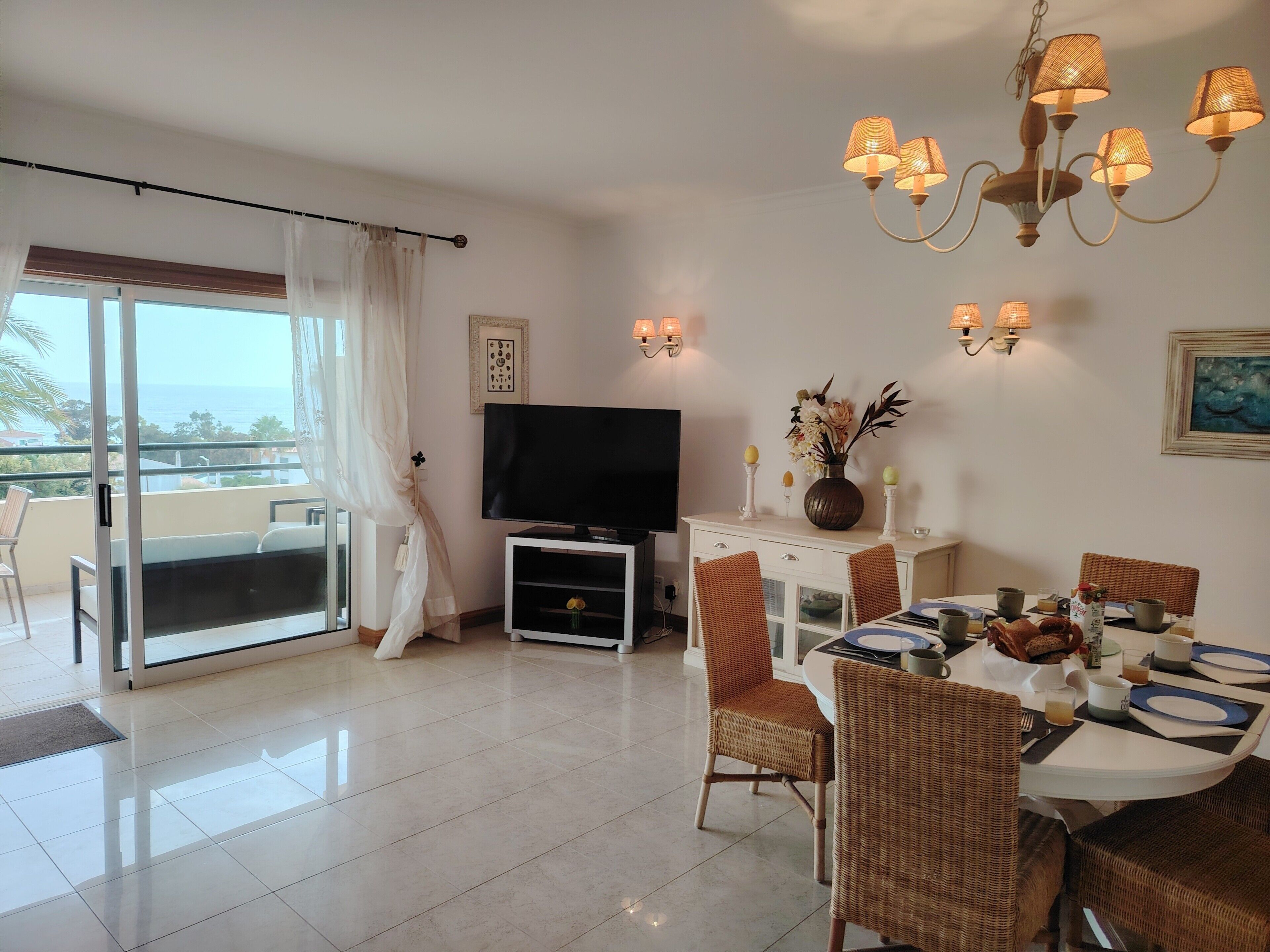 A dining area with a table set for two, a television, and a balcony with curtains.