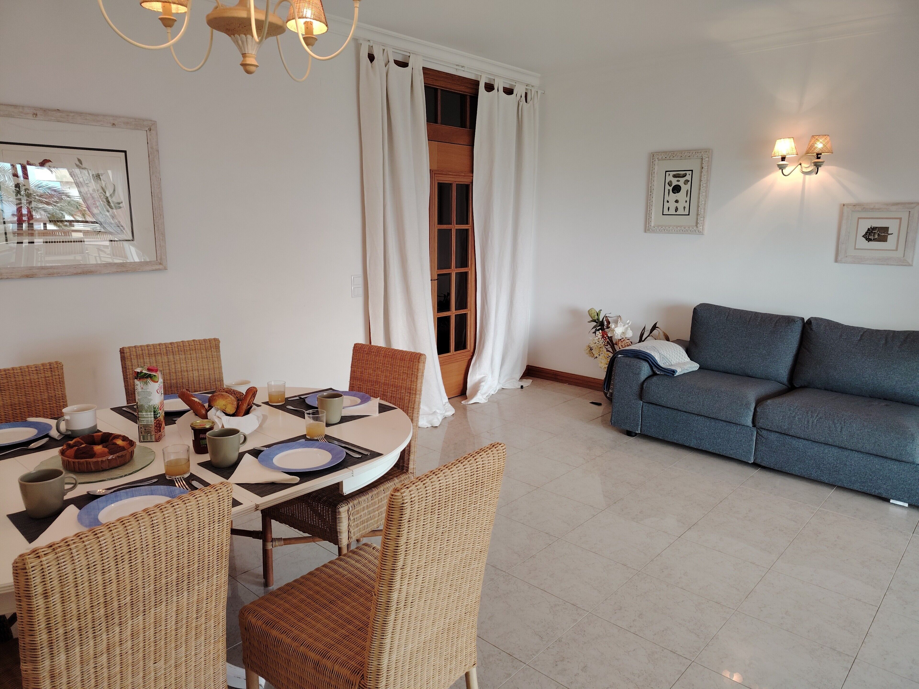 A dining area with wicker chairs, a table set for two, and a blue sofa in the background.