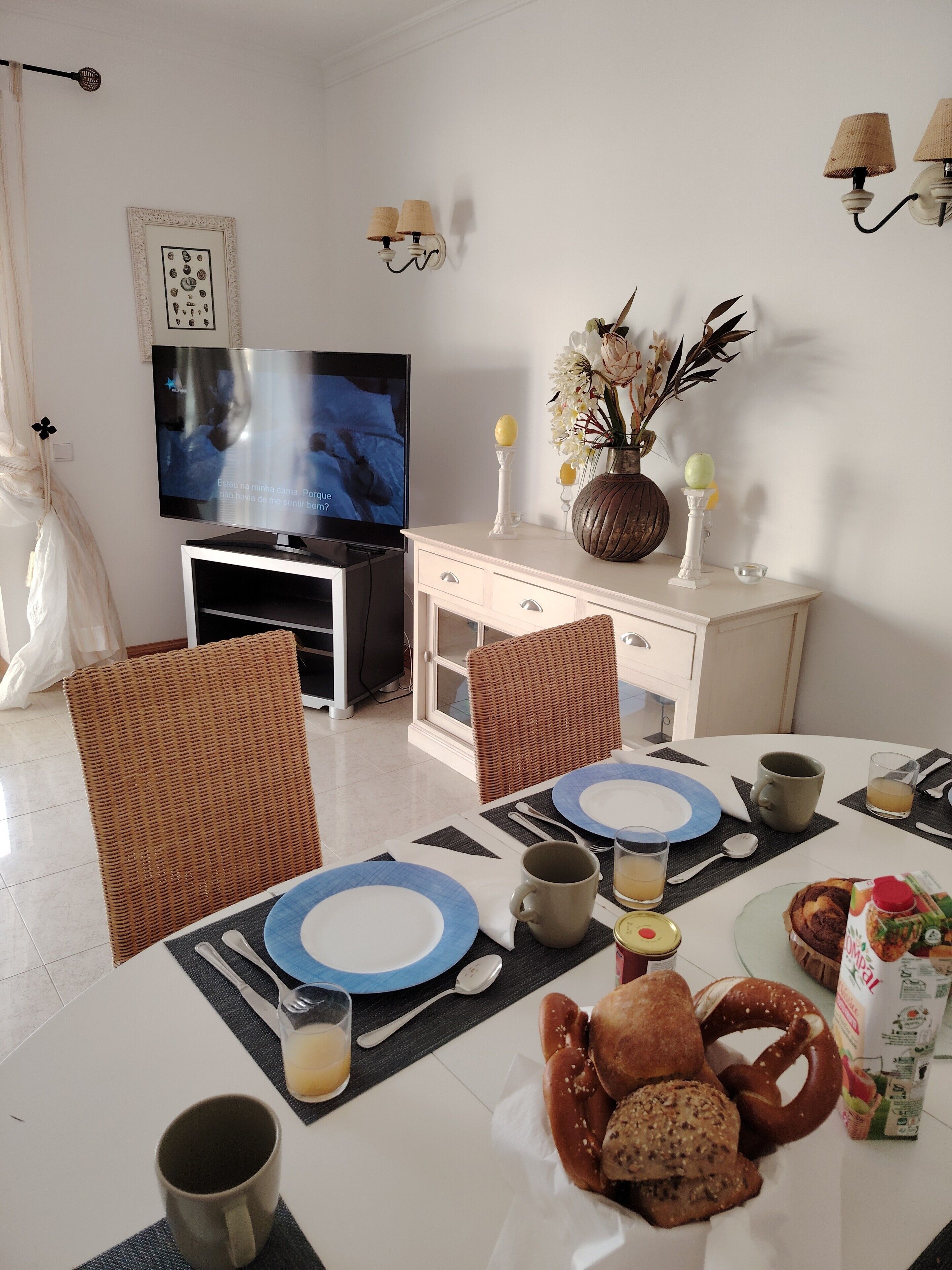 A dining area with a television, wicker chairs, a white table with blue plates, and a variety of food items.