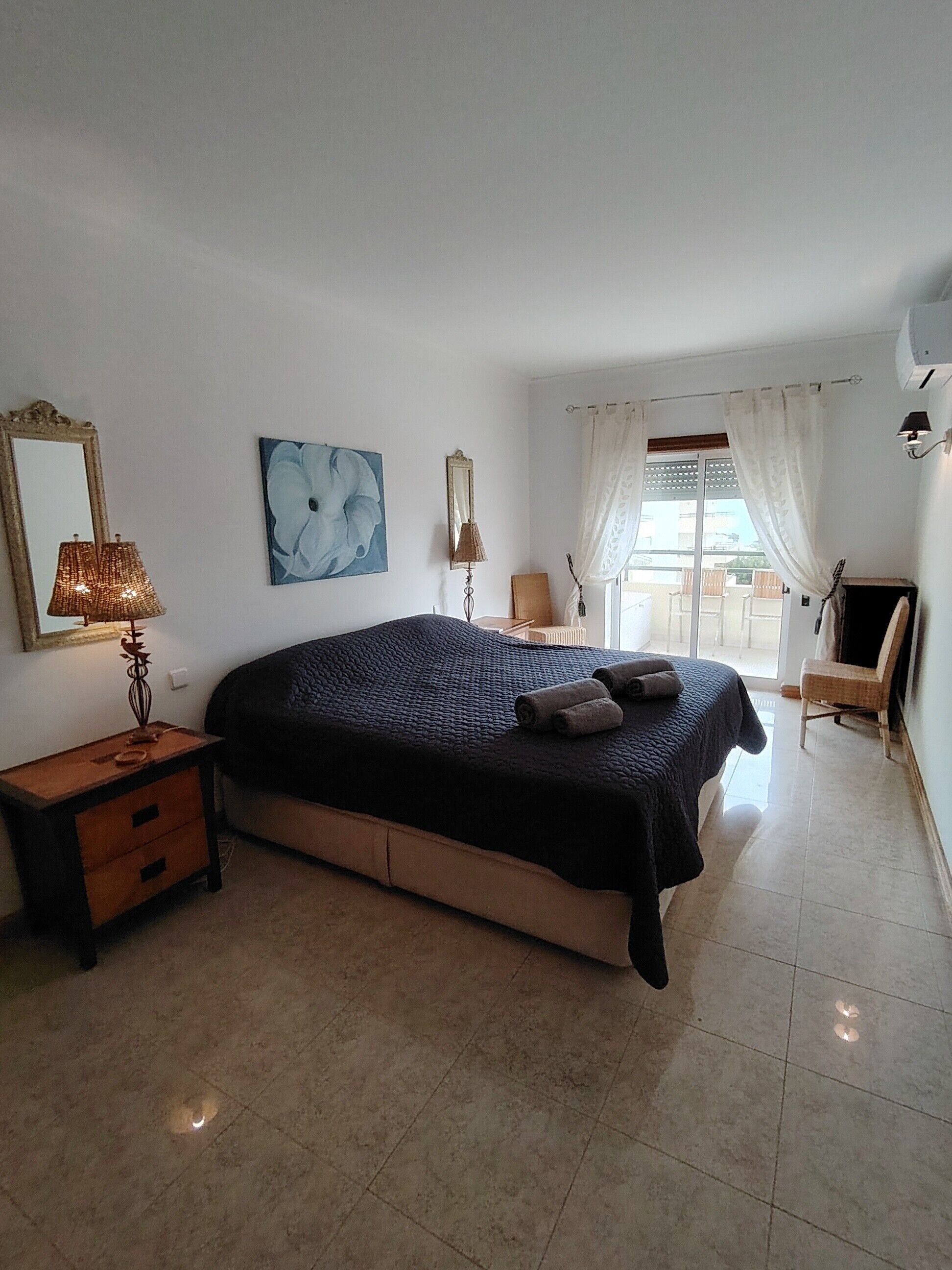 A bedroom with a bed, a nightstand, a lamp, a painting, a chair, and a window with curtains.