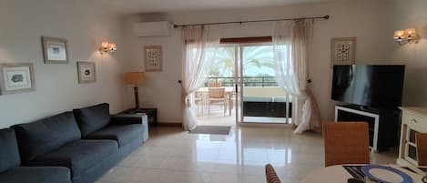 A living room with a sofa, a dining table set for two, a television, and a balcony with curtains.