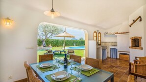 A dining area with a table set for two, a kitchen with a brick oven, and a view of a swimming pool and garden. - Peaceful Rural Haven With Pool (Pollença)