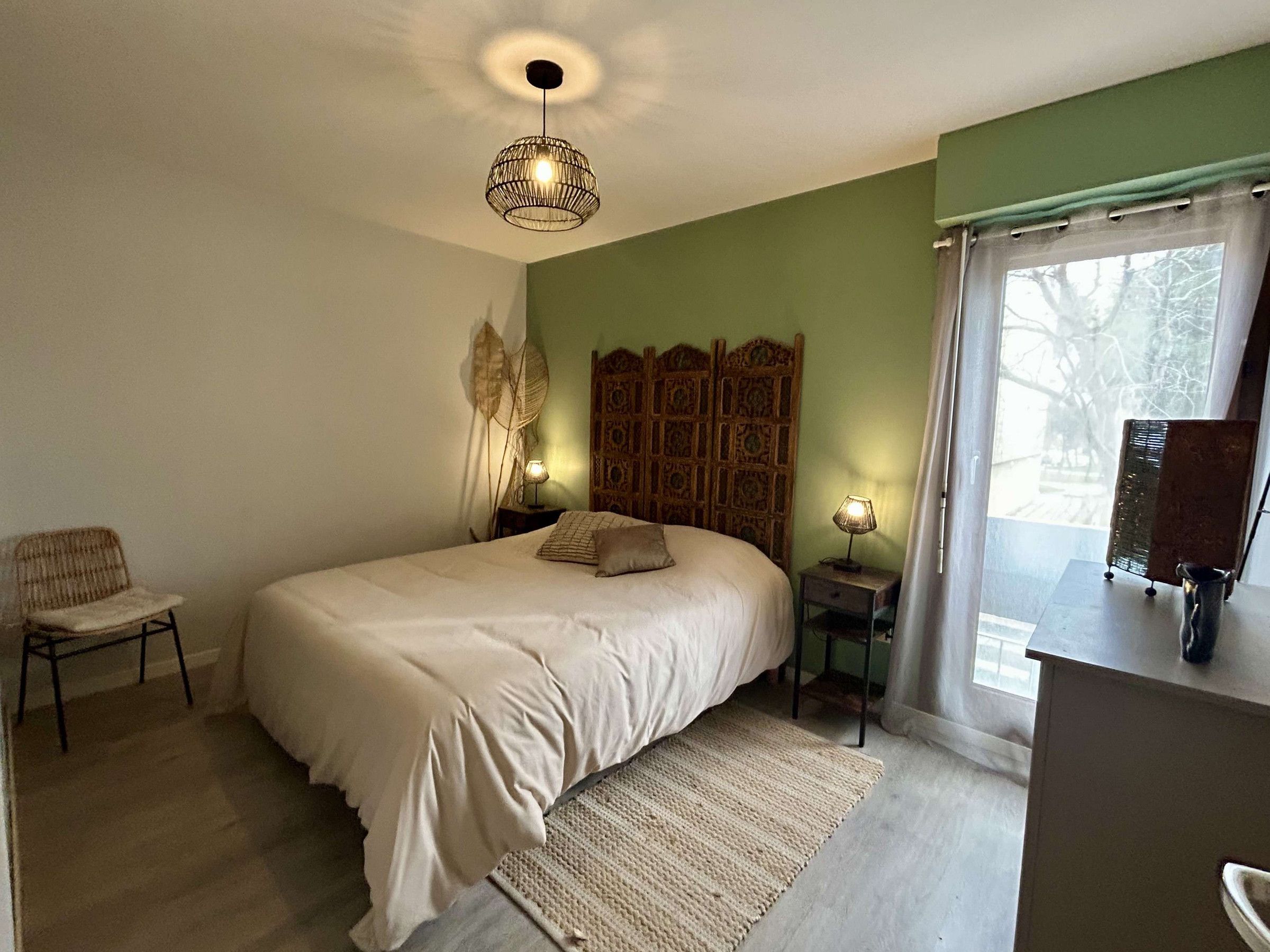 Quarto com cama grande, uma cadeira, mesa de cabeceira com abajur, cabeceira decorativa e vista para o exterior pela janela.