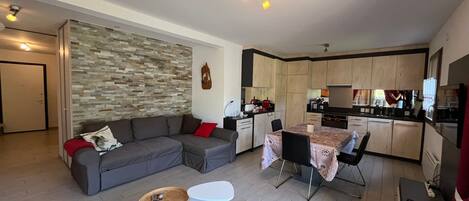 A modern living space with a grey sectional sofa, a dining table with chairs, a kitchen area, and a red rug.