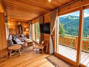 A wooden cabin interior with a sofa, table, chairs, a TV, and a balcony with a forest view. - Spacious apartment, mountain view - 4 guests (La Clusaz)