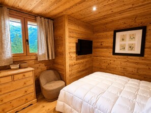 A wooden cabin room with a bed, a chair, a dresser, a window with curtains, and a framed botanical print on the wall. - Spacious apartment, mountain view - 4 guests (La Clusaz)