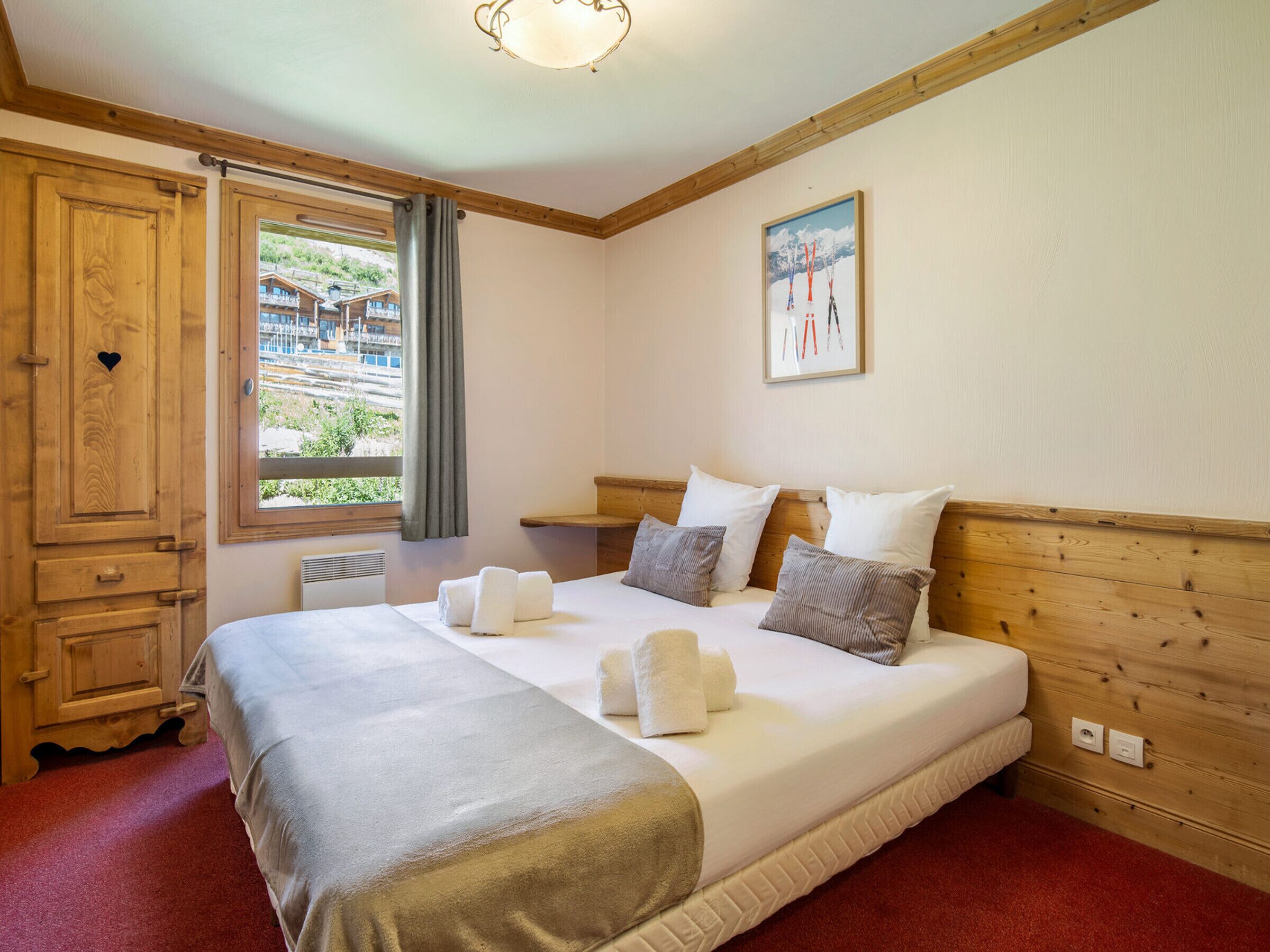 A hotel room with a large bed, wooden headboard, a window with curtains, a wooden wardrobe, and a painting on the wall.