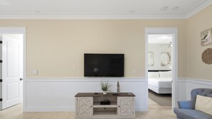 Uma sala de estar com televisão de tela plana na parede, mesa lateral de madeira com gavetas, um sofá azul e uma cama ao fundo.