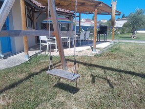 A wooden swing set with a bench, white plastic chairs, and a striped umbrella under a wooden pergola.
