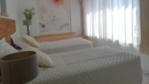 Ett hotellrum med två sängar, ett stort blommotiv på väggen, en krukväxt och ett fönster med tunna, genomskinliga gardiner.