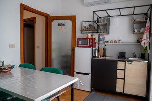Eine kompakte Küche mit Kühlschrank, Mikrowelle und Kaffeemaschine. Ein Esstisch mit Stühlen und eine kleine Arbeitsfläche mit Regalen vorhanden.