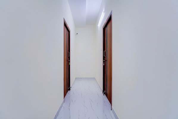 A corridor with two wooden doors on either side, a white tiled floor, and a ceiling with a fluorescent light fixture.