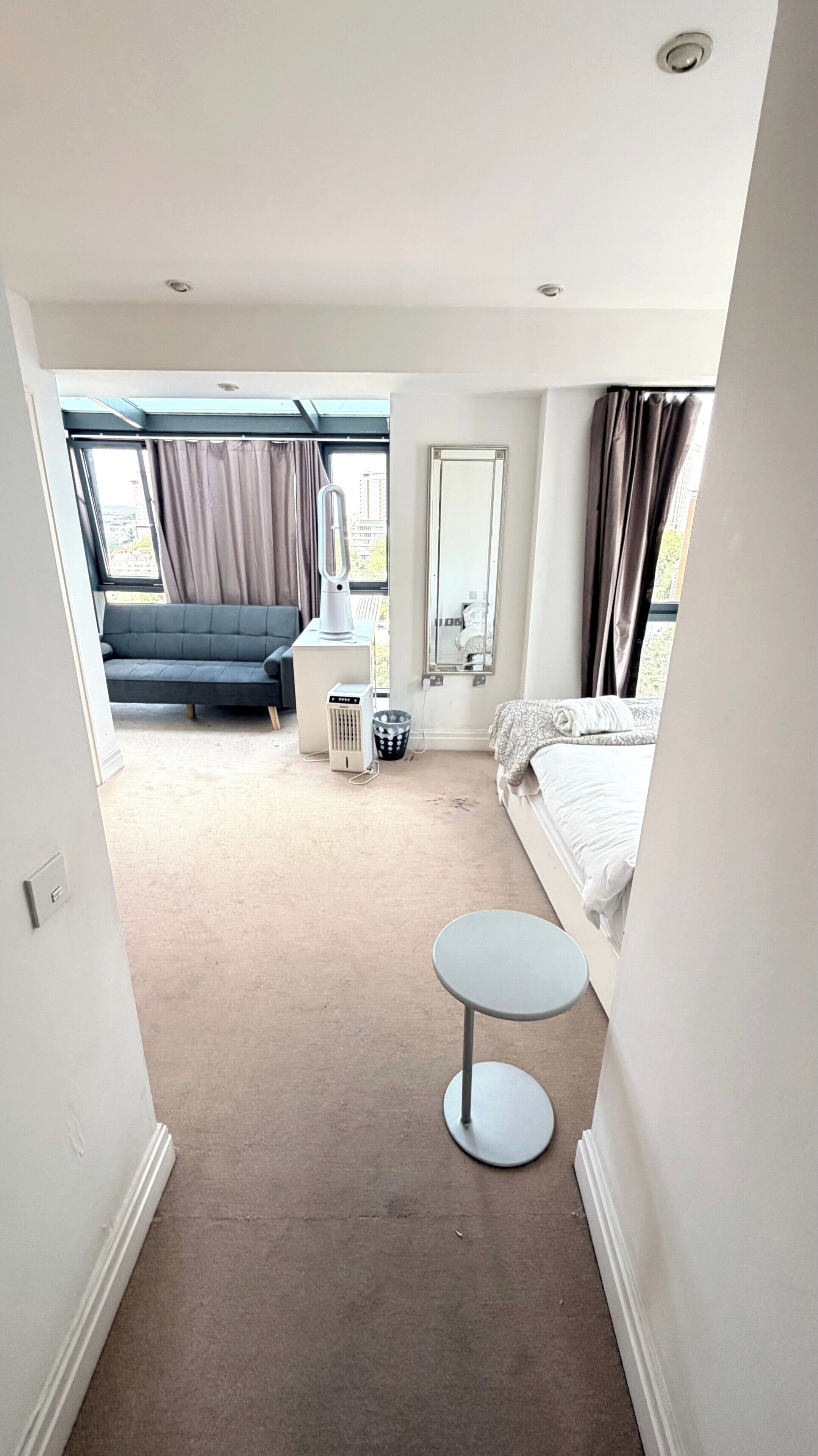 A hotel room with a bed, a small round table, a sofa, a heater, a trash bin, and a window with curtains.