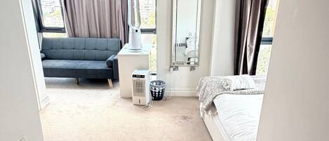 A hotel room with a bed, a small round table, a sofa, a heater, a trash bin, and a window with curtains.