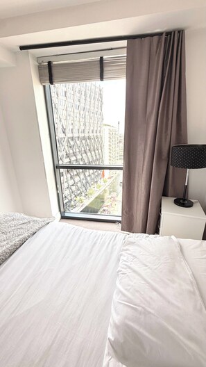 Ein Hotelzimmer mit einem großen Bett, einem Nachttisch mit Lampe und Blick auf eine Stadtlandschaft durch ein Fenster mit Jalousien.