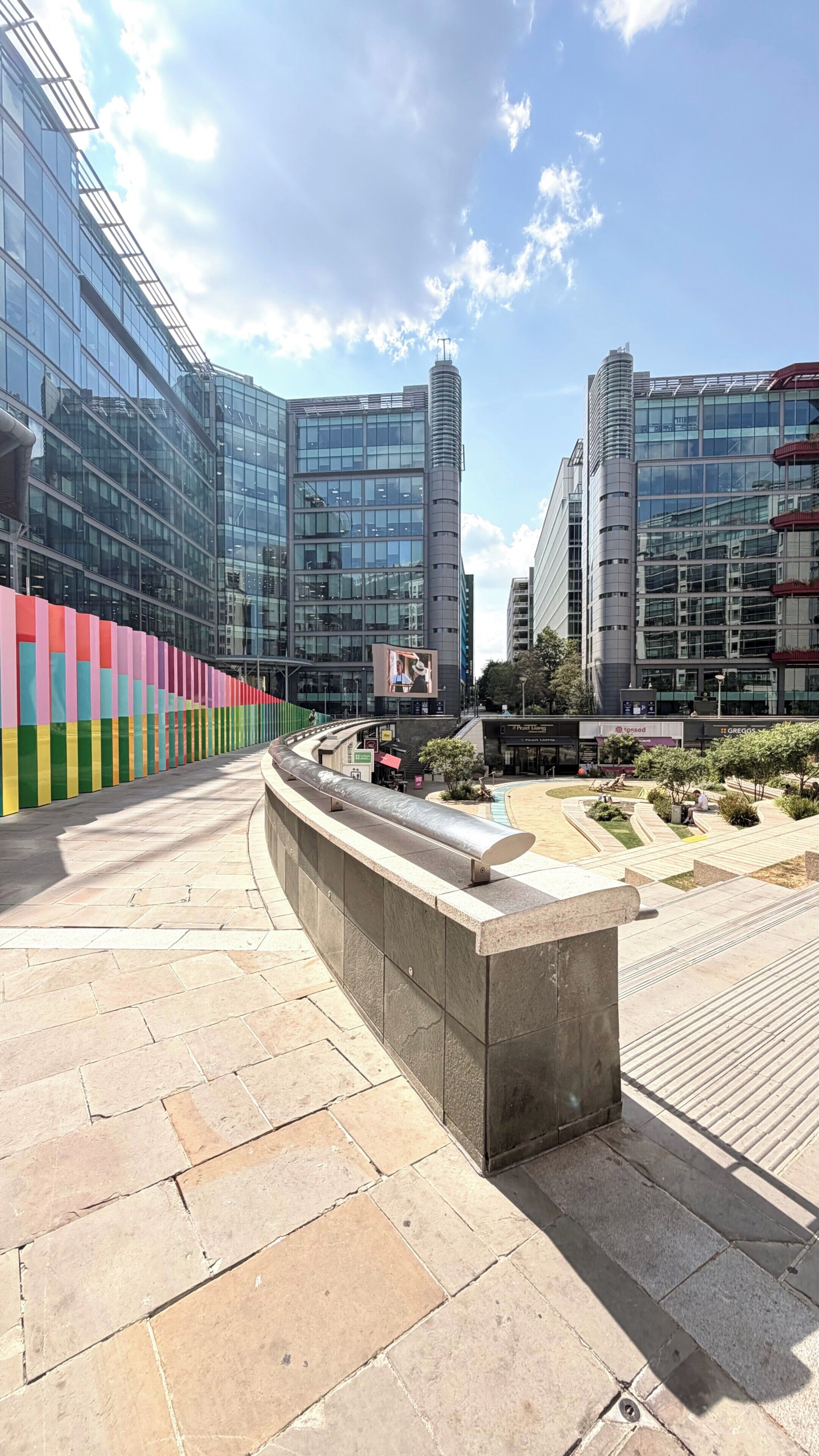 A modern urban plaza with glass buildings, a curved seating area, and a colorful fence.