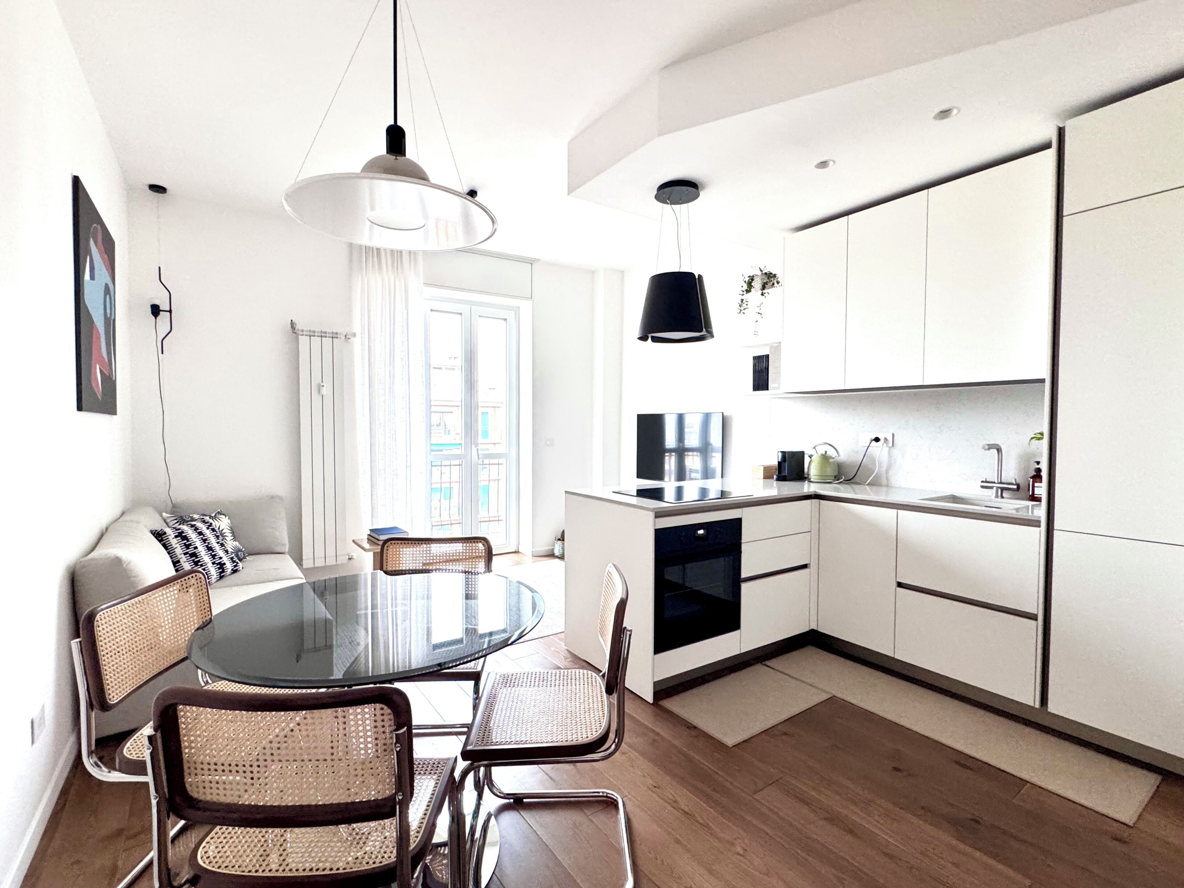 A modern kitchen with white cabinets, a dining area with a glass table, and a black pendant light.