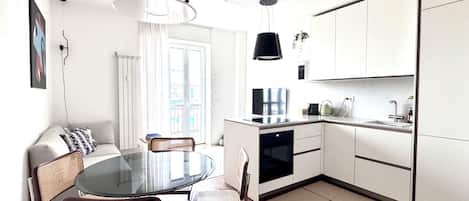 Una cocina moderna con armarios blancos, un comedor con mesa de vidrio y una lámpara colgante negra.