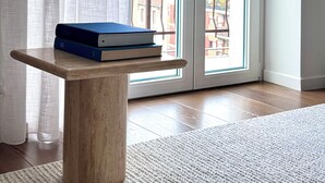 Een kamer met een houten tafel, een raam met uitzicht op gebouwen en een wit gordijn.
