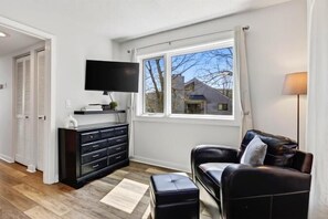 Una sala de estar moderna con un sofá negro, un televisor de pantalla plana montado sobre un mueble negro, y una ventana con vistas al exterior.