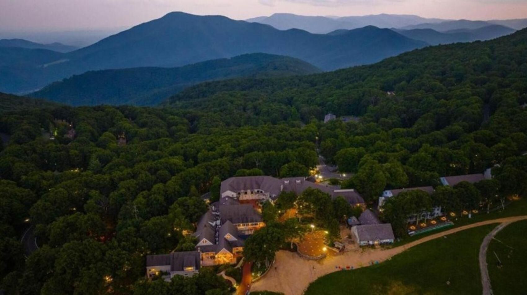 An aerial view of a residential area with multiple houses surrounded by dense forest, featuring a central building with a prominent roof and a well-lit driveway.
