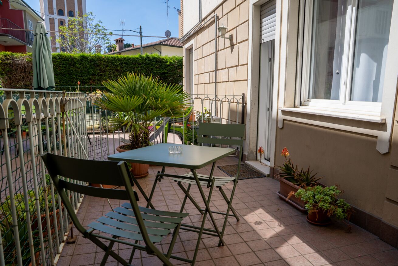 Una terrazza esterna con tavolo e sedie, circondata da piante in vaso e una recinzione metallica.